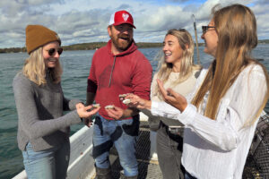 Four people smiling and holding out oysters to eat on a boat