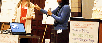 two women, one with a microphone, standing among flipcharts