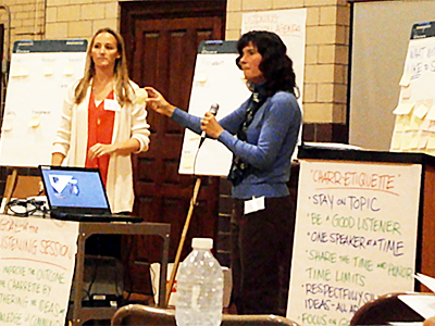 two women, one with a microphone, standing among flipcharts