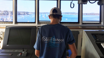 a teenager at the helm of a boat wearing a Maine Ocean School t-shirt