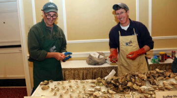 Two smiling men shucking shellfish behind a table of scallops and oysters on the half-shell