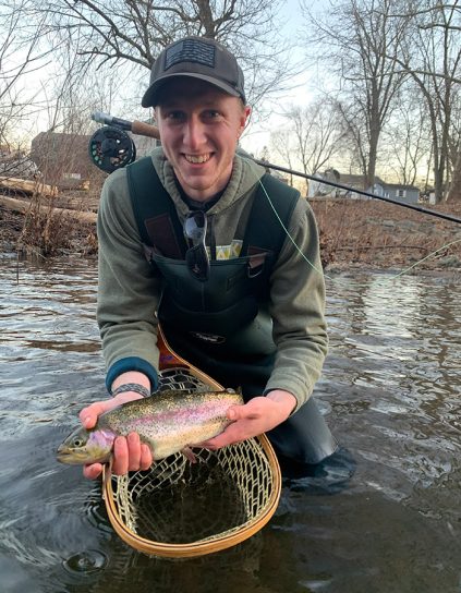 Dylan Garrabrant knee deep in the water with a flyfishing rod, holding a net and a fish