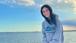 Aimee Whitman with backdrop of the ocean wearing a UMaine lacrosse pullover
