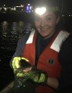 Andrea Casey at night in a headlamp holding a fish in gloved hands water in the background