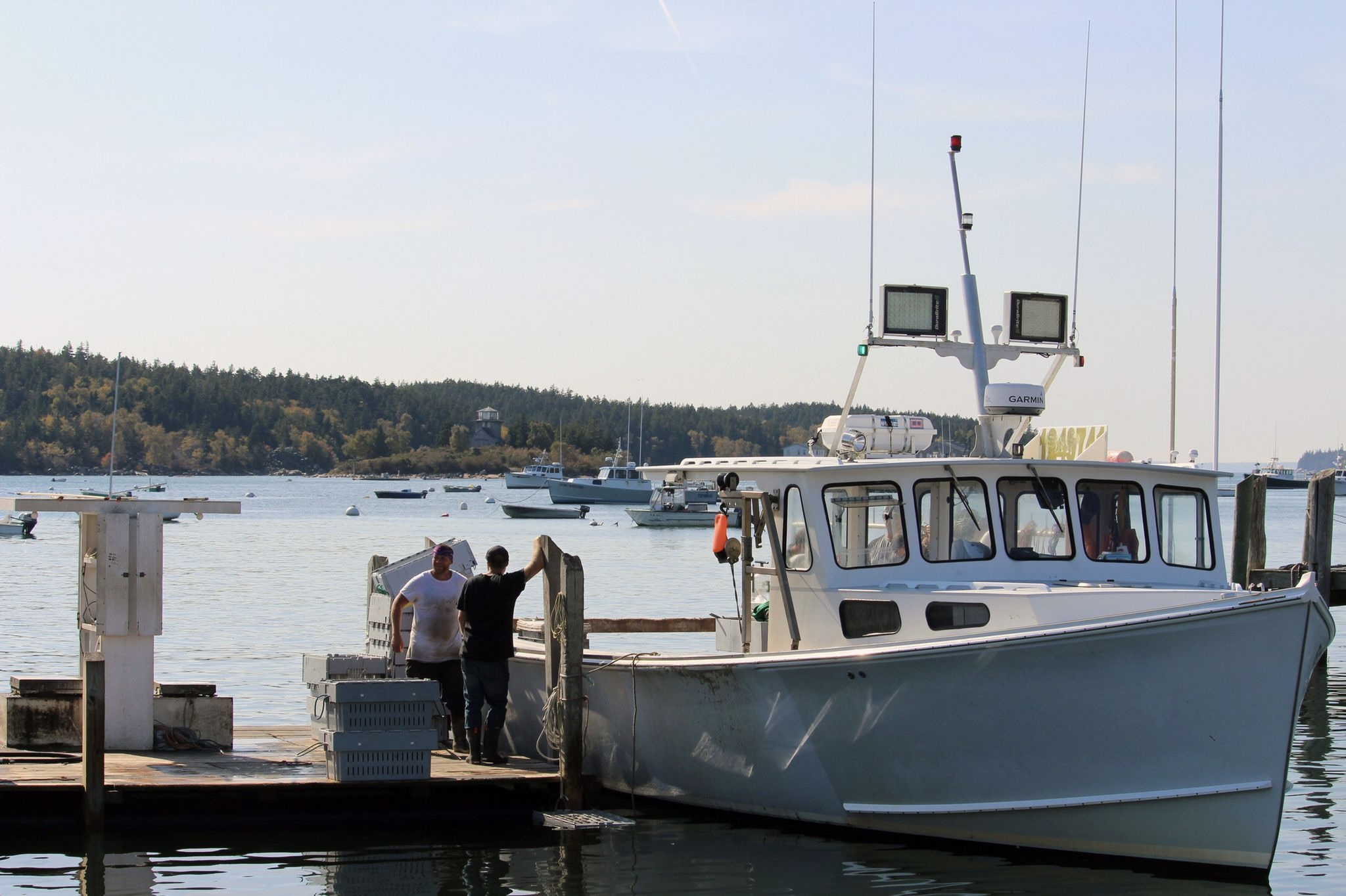 2 people stand on dock next to a lobster boat in the water.