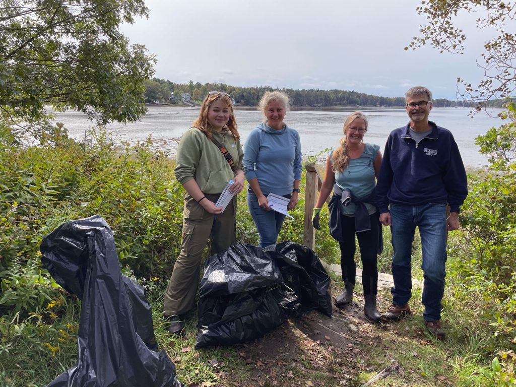 Four people stand on the shoreline with trash bags at a Maine coastal cleanup event. 