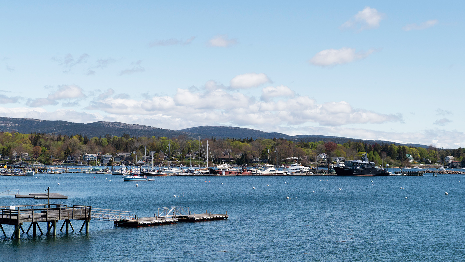 A wide landscape of Southwest Harbor. There is a wharf in the foreground, many boats in the harbor, and the mountains of Acadia National Park in the distance.
