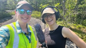 A two-person selfie, both people smiling, the one on the left in a yellow reflective vest and sunglasses, the other wearing headphones, in front of a wooded background.