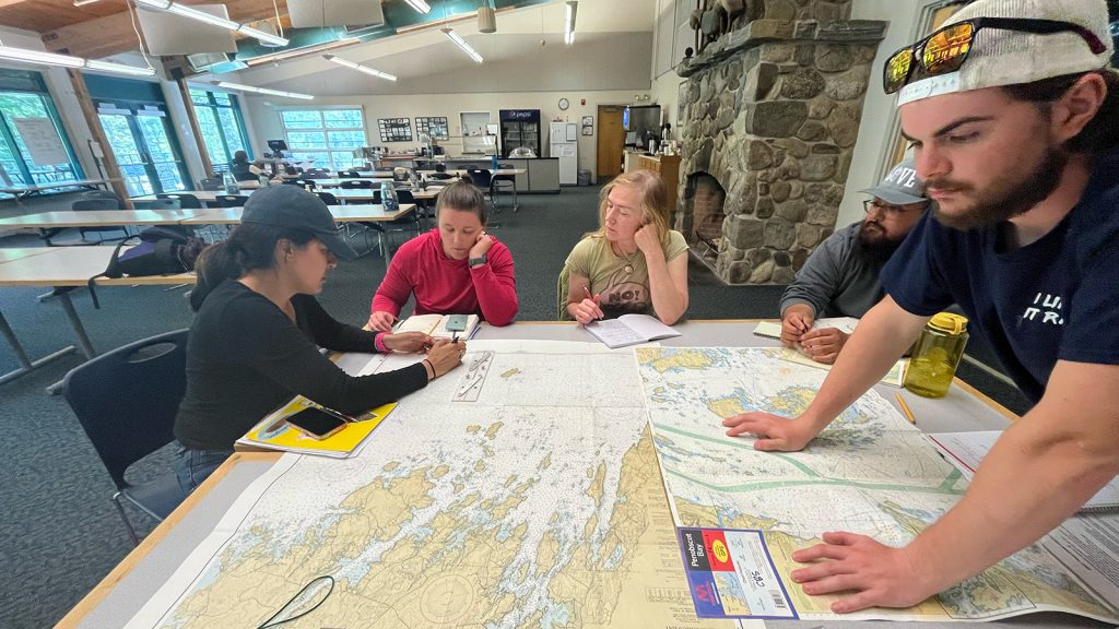 Several people stand and sit around a table covered in nautical charts, compasses, and other navigation gear.