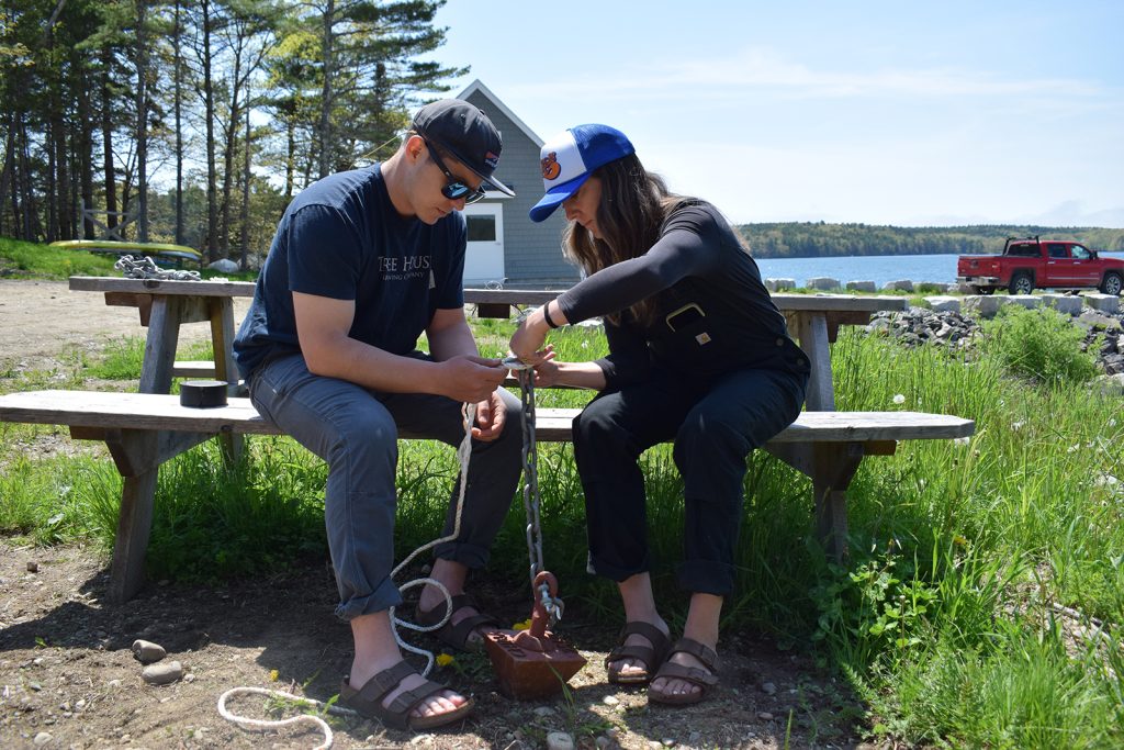 A man and a woman sit together on a picnic table bench holding an anchor, chain, and some rope.