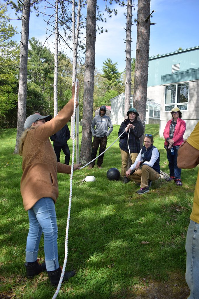 A group of people outside stand around a woman holding up a rope and a piece of pipe.