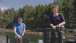 A woman and a man standing by the water holding black plastic shellfish bags