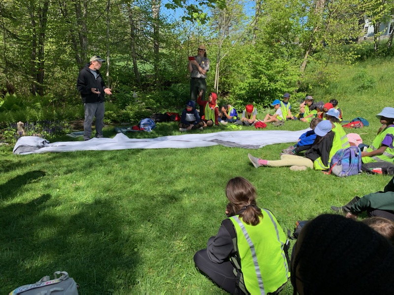 A man in a hoodie is standing by a long white, cylindrical fishing net with a small mesh. Children sit around him listening intently.