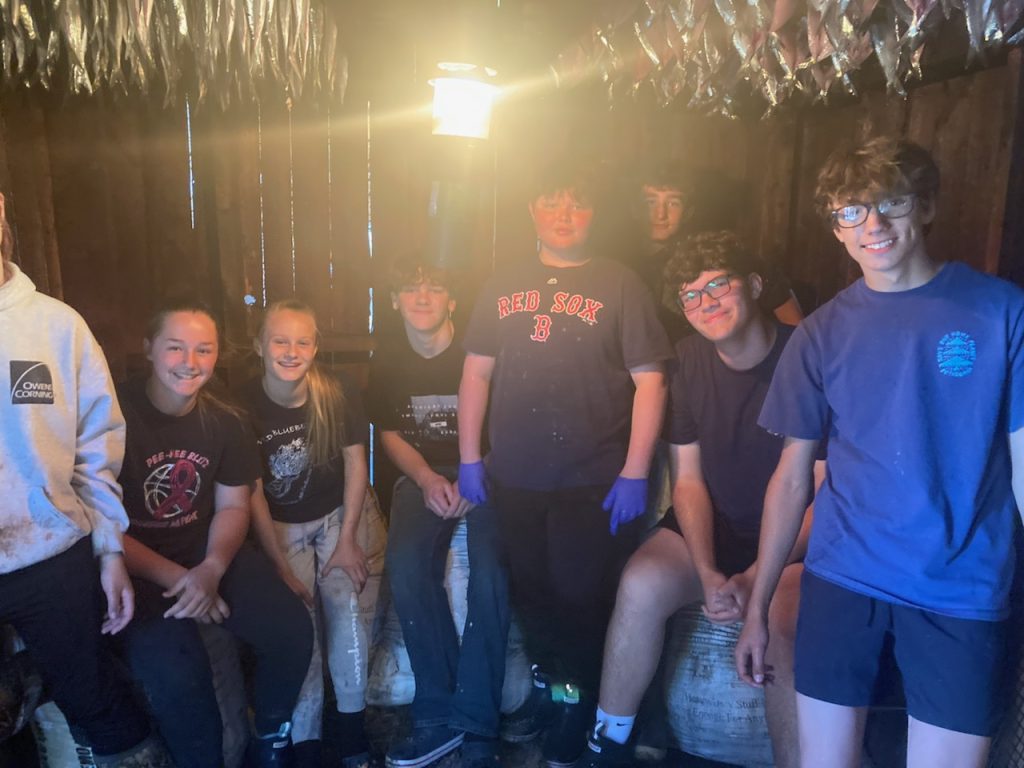 Students stand in a half circle in the smokehouse. Fish are hanging from sticks on the ceiling and a yellow lamp glows, hanging from the ceiling.