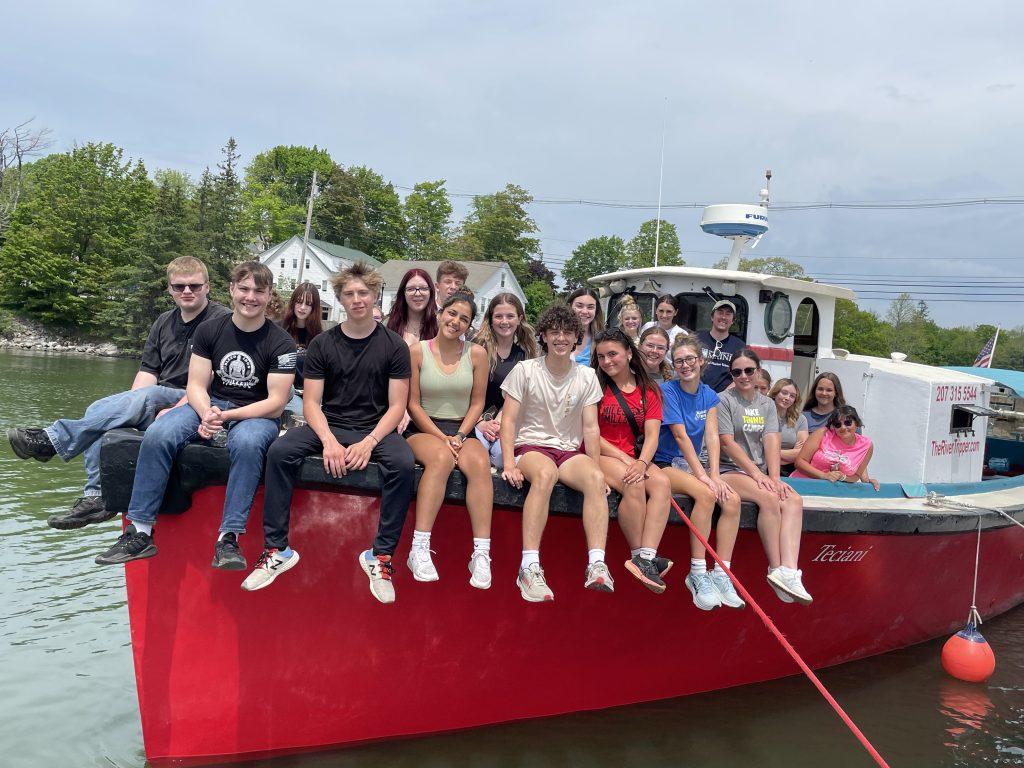 Students sit on the bow of a red boat.