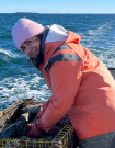 Michelle in a pink cap and orange jacket with the ocean behind her