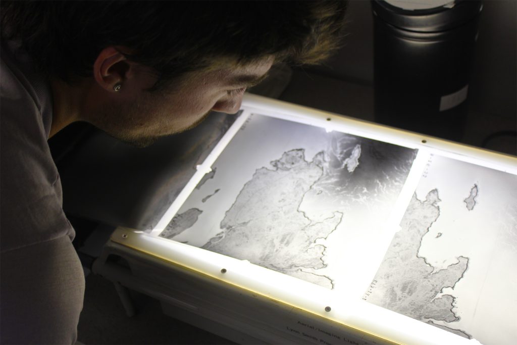 A person holds their face to a glowing table to look at a negative photograph. The black and white image is a snapshot of a craggy peninsula.