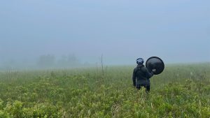 A figure standing in a field of vegetation with a round listening device looking into the blue fog