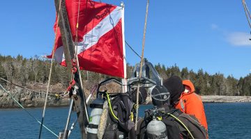 Facing the back left side of the boat, two divers are putting on gear for a dive. Extra gear is laid out behind them on a covered rubber hot tub for easy access. A dive flag on the left back corner of the boat is moving in the wind. The safety diver wearing an orange raincoat is next to the flag in front of the divers. Water surrounding the boat is Frenchman Bay and an island coastline.