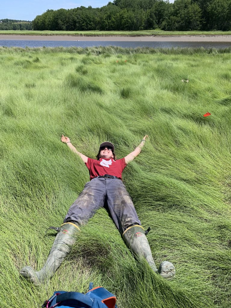 Student lays with their arms above their head taking a rest on top of a grassy tidal marsh.
