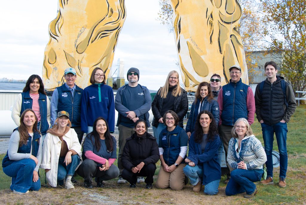 sixteen people standing and kneeling in front of the yellow painted legs of a lobsterman statue near the ocean